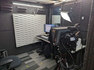 Interior of the recording booth with black felt walls, standing desk, camera with teleprompter, lights, and white decorative wave pattern wall.